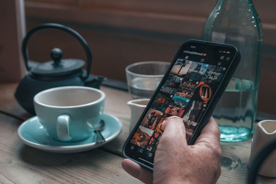 A person holds a smartphone browsing Instagram next to a teacup and teapot on a wooden table.